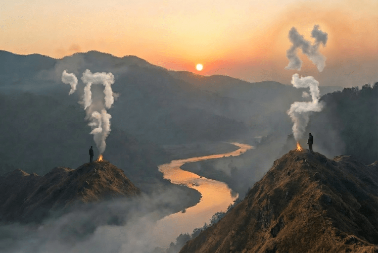 Silhouetted people by a campfire overlooking a foggy mountain valley during sunrise.